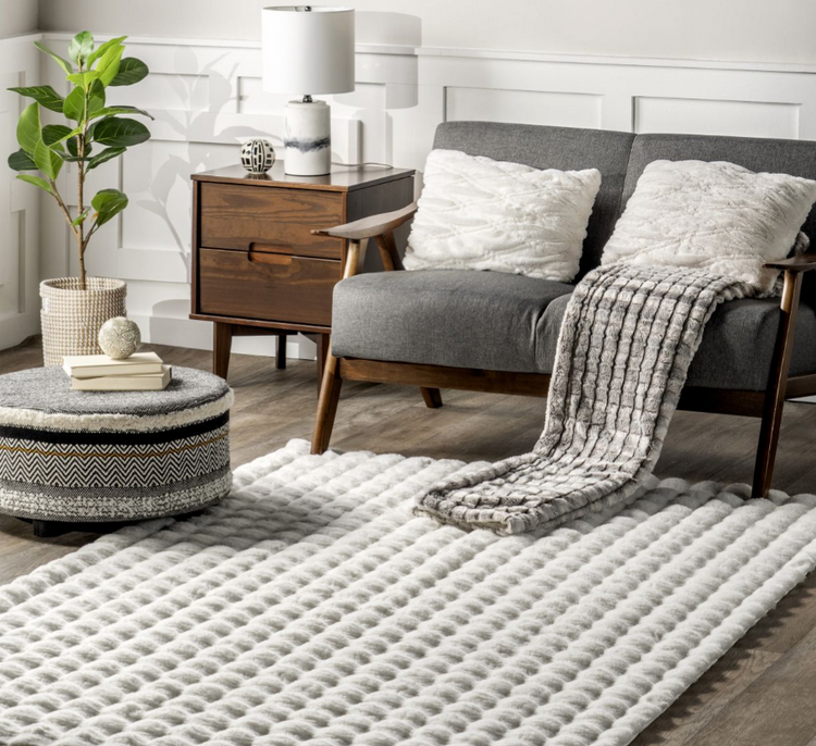 Modern living room with gray sofa, white textured pillows, cozy gray throw, round patterned ottoman, wooden side table, lamp, and indoor plant