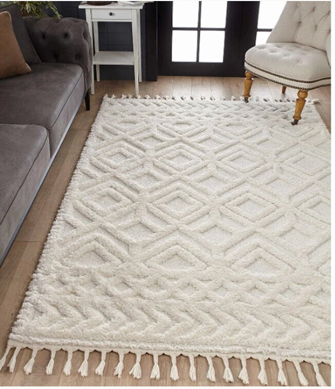 White textured geometric patterned area rug with fringe on hardwood floor between gray sofa and beige upholstered chair