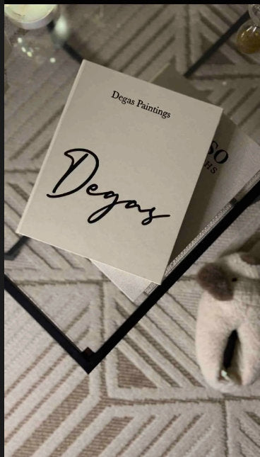 Open book titled 'Degas Paintings' on glass coffee table with geometric rug and fuzzy slippers