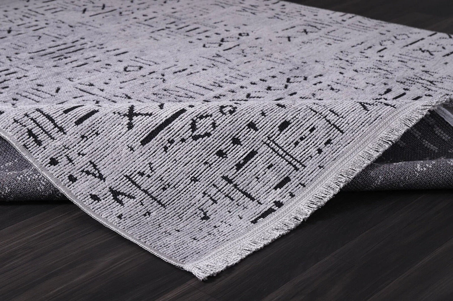Light gray rug with abstract black pattern on dark wooden floor, corner lifted showing weave