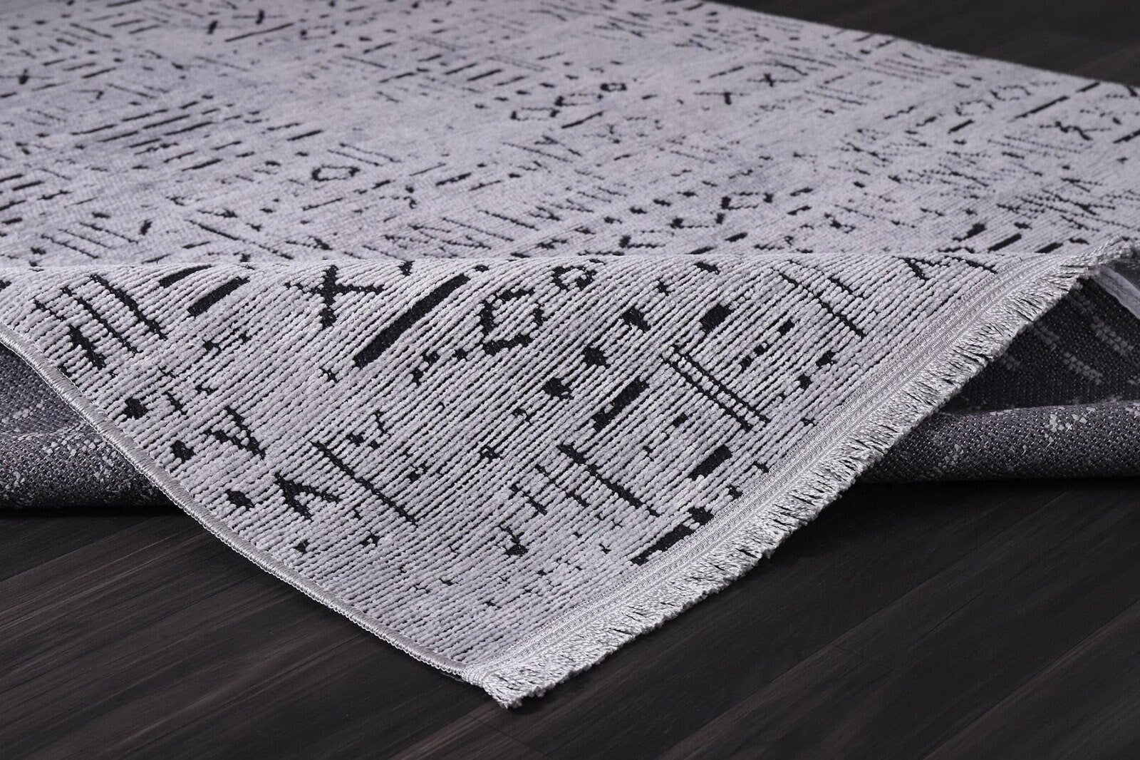 Light gray rug with abstract black pattern on dark wooden floor, corner lifted showing weave
