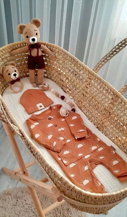 Woven baby bassinet with orange sun-patterned onesie, matching hat, rattle, pacifier, and brown crocheted teddy bear toy