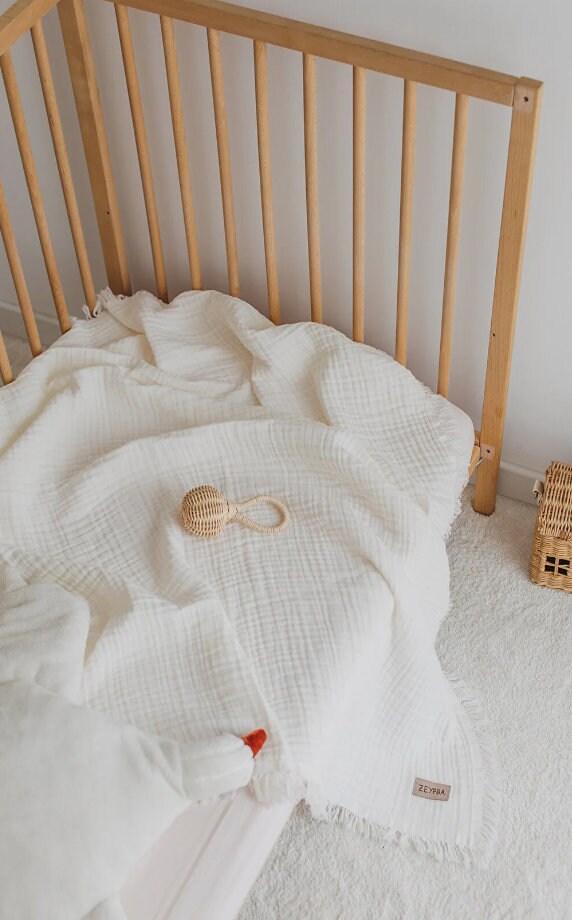 Minimalist wooden baby crib with soft white muslin blanket and wicker rattle on carpeted floor